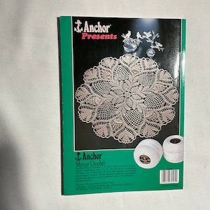 May include: A book cover featuring a white crochet doily with a floral design. The book cover is black with the text "Anchor Presents" and "Anchor Mercer Crochet" in red and white. The book cover also features a white ball of yarn.