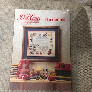 May include: A cross-stitch leaflet with a colorful handprint design and a poem about a child's growing hand. The leaflet is titled "Handprints" and is from J&P Coats. The poem reads: "You sometimes get discouraged because I am so small and always leave my fingerprints on furniture and walls. But every day I'm growing and all those fingerprints will surely fade away just so you can recall how my little hand looked when I was very small."