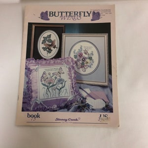 May include: A book cover titled "Butterfly Wings" featuring cross-stitch designs. The cover displays framed butterfly and floral patterns, including a pillow with the text "Follow Your Dreams in Butterfly Wings."