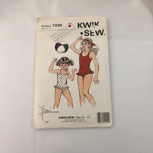 Könnte beinhalten: Ein Schnittmuster für zwei Kinder-Badeanzüge. Das Schnittmuster ist von Kwik Sew Pattern Co., Inc. und ist für die Größen 4-5-6-7. Das Schnittmuster zeigt zwei Mädchen, die die Badeanzüge tragen, eines in einem rot-weiß gepunkteten Badeanzug und das andere in einem rosa-weiß gemusterten Badeanzug mit Herzen.