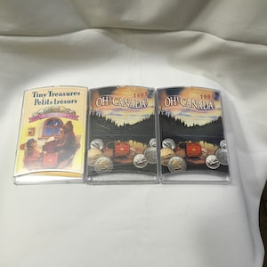 Op de afbeelding: Drie transparante plastic koffers met sets Canadese munten. De eerste koffer heeft een kleurrijke illustratie van een beer die een boek leest met de tekst "Tiny Treasures Petits trésors 1999 - 2009". De andere twee koffers hebben een zwart-witte illustratie van een bos met de tekst "Oh! Canada! 1999" en "Oh! Canada! 1998".