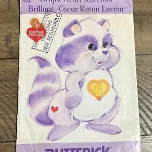 May include: A vintage sewing pattern featuring a cartoon raccoon character with purple and white fur, a heart-shaped lightbulb on its chest, and a heart on its rear. The pattern is labeled "Bright Heart Raccoon" and "Butterick."