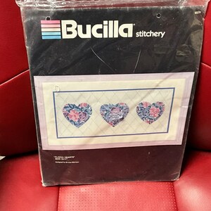 May include: A Bucilla stitchery kit featuring a cross-stitch design of three floral hearts. The kit is packaged in a clear plastic bag with a black and white label that reads "Bucilla stitchery" and "Floral Hearts".