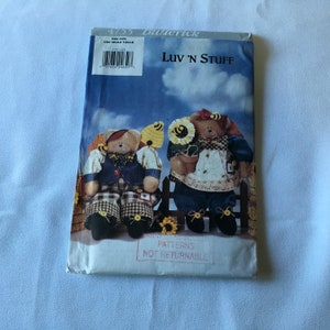 May include: A sewing pattern for two teddy bears, one wearing a blue shirt and plaid trousers, and the other wearing a blue dress and plaid skirt. The pattern is titled "Luv 'N Stuff" and is from Butterick.
