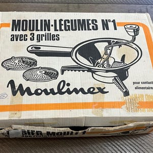 Peut inclure: Boîte de moulin à légumes Moulinex vintage, avec texte en français. La boîte blanche et orange présente une illustration en noir et blanc du moulin et de trois grilles. La boîte est légèrement usée.