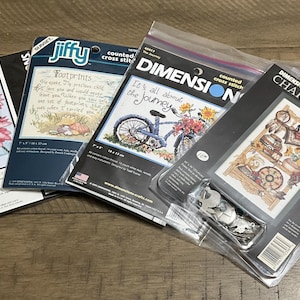 May include: Several counted cross stitch kits featuring designs of butterflies, flowers, a bicycle, and vintage charts. The kits are packaged in plastic and cardboard. Measurements are in cm. Various brand names are visible.