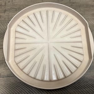 May include: A circular, light beige tray featuring a raised, sunburst-inspired pattern of radiating lines that extend from the center toward the outer rim. The tray has a smooth, matte finish and a slightly elevated edge, creating a structured design.