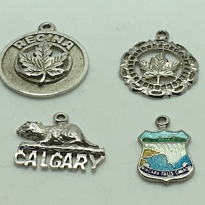 Op de afbeelding: Vier zilveren bedels met verschillende ontwerpen. De eerste bedel is rond en heeft een zilveren esdoornblad met het woord "REGINA" aan de rand. De tweede bedel is een zilveren esdoornblad met een decoratieve rand. De derde bedel is een zilveren bever met het woord "CALGARY" eronder. De vierde bedel is een zilveren schild met een blauw en wit emaille ontwerp van Niagara Falls en de woorden "NIAGARA FALLS CANADA".