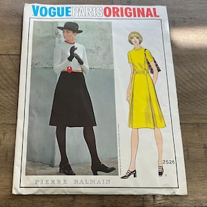 May include: Vogue Paris Original sewing pattern featuring two fashion illustrations. One shows a woman in a black skirt, white top, and hat. The other shows a woman in a yellow dress. The pattern is by Pierre Balmain.