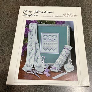 May include: A printed pattern booklet titled Lilac Chatelaine Sampler by The Victoria Sampler. The cover displays a cross-stitch project featuring a long light blue ribbon, a framed sampler, a small lavender-themed sachet, and heart-shaped ornaments.