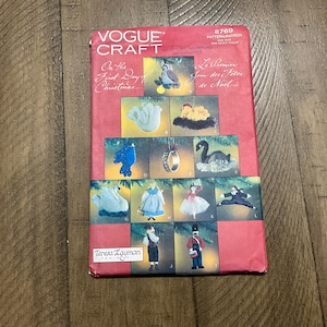 May include: A vintage Vogue Craft pattern packet with a red cover. The cover features illustrations of Christmas ornaments, including a swan, owl, and ring. The text reads "On the First Day of Christmas" and "Le Premier Jour des Fêtes de Noël."