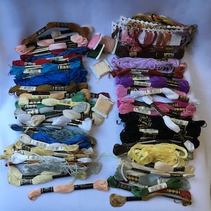 May include: A large collection of embroidery floss spools in various colors, including white, black, red, blue, green, yellow, and brown. The spools are arranged in rows and columns, and some are stacked on top of each other. The spools are labeled with numbers and letters.