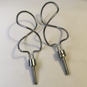 May include: Two stainless steel mixer beaters with a distinctive looped design. The beaters have a spiral shape at the top and a cylindrical base, perfect for mixing ingredients. The image is taken on a white background.