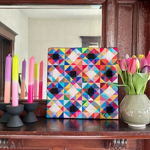 May include: A colorful geometric quilt pattern artwork with black squares and a variety of colors, including pink, blue, green, yellow, and red. The artwork is framed and hanging on a wall above a wooden shelf with candlesticks and a vase of pink tulips.