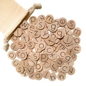 May include: A set of wooden letter tiles in a natural wood color with the letters of the alphabet printed on them. The tiles are scattered on a white surface and a small canvas bag is in the upper left corner of the image.