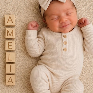 May include: A baby wearing a beige ribbed long-sleeve romper with three small wooden buttons at the neckline and a matching beige bow headband. Beside the baby are seven light wooden blocks arranged vertically, each featuring the letters A, M, E, L, I, A.