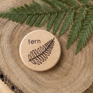 May include: A circular light-colored wooden disc featuring a dark brown laser-engraved illustration of a fern leaf and the word fern printed in lowercase letters. The wood grain is visible on the surface of the smooth, flat, round object.