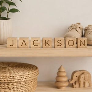 May include: Seven light-colored wooden blocks arranged in a row on a shelf, each featuring a single engraved letter. Together, the blocks spell out the name JACKSON in a clean, sans-serif font, with visible wood grain texture on each cube.