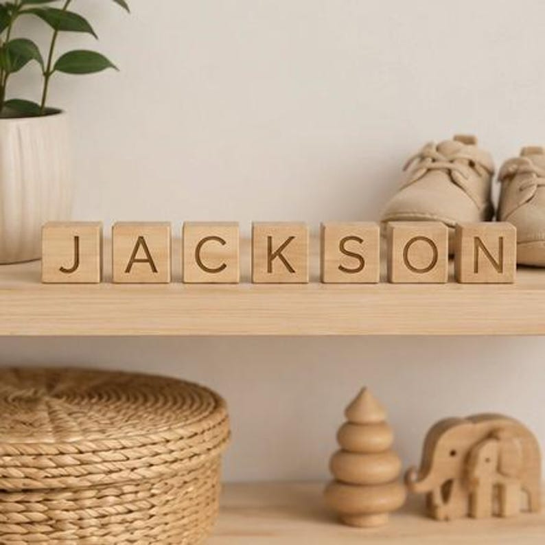 May include: Seven light-colored wooden blocks arranged in a row on a shelf, each featuring a single engraved letter. Together, the blocks spell out the name JACKSON in a clean, sans-serif font, with visible wood grain texture on each cube.