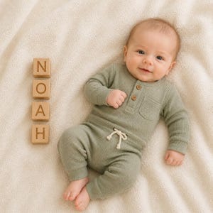 May include: A sage green long-sleeve ribbed romper with a three-button henley neckline, a small chest pocket, and a drawstring waist. Beside the romper are four wooden blocks stacked vertically, each featuring one letter to spell out the name NOAH.