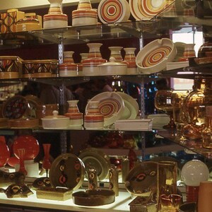 May include: A display of vintage ceramic and glass items, including vases, bowls, and serving dishes. The items feature retro designs with orange, brown, and white color schemes. The shelves also hold red and amber-colored glass pitchers and decorative objects.