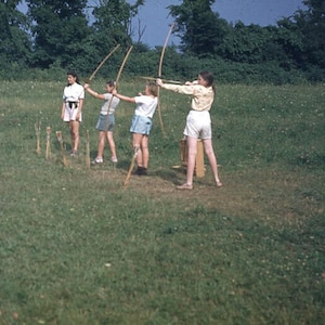 May include: Four people are practicing archery in a grassy field. They are aiming at targets made of wooden stakes. The archers are wearing shorts and short-sleeved shirts. The background features a line of trees under a blue sky.