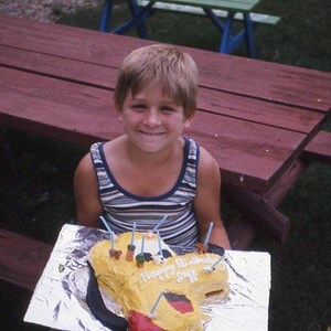 May include: A young person smiles while holding a yellow airplane-shaped birthday cake with the words "Happy Birthday, Guys" written on it. The person wears a striped tank top and sits in front of a red picnic table.
