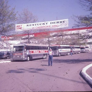 May include: A vintage photograph shows a row of silver and red buses parked on a paved area. The buses have the words "Michigan Railways" on the side. A banner overhead reads "Kentucky Derby at Ladbroke-DRC". A person stands near the buses.