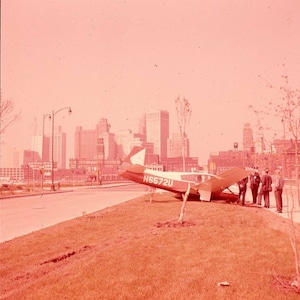 May include: A small red and white airplane with the registration number N6572U is resting on a grassy area next to a road. The plane is angled, with its wing touching the ground. Several people are standing nearby, and a city skyline is in the background.