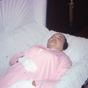 May include: A person in a pink dress and white gloves lies in a white casket. The casket is lined with white fabric. The background is dark.
