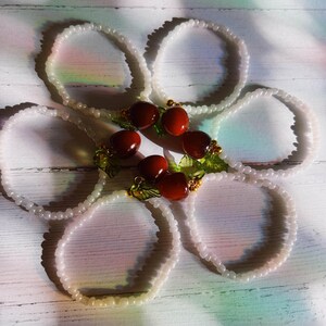 May include: Five white beaded bracelets with red and green cherry charms. The charms have gold accents.