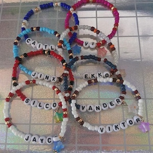 Puede incluir: Una colección de pulseras de cuentas de varios colores, incluyendo azul, rojo, blanco y rosa. Algunas pulseras presentan nombres deletreados con cuentas de letras. Cada pulsera tiene un dije, como una estrella o un colgante colorido.