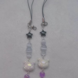 May include: Two matching phone charms. Each charm features a black star, light blue beads, a white bear, and a pink heart. The charms are connected to a black cord with a silver clasp.