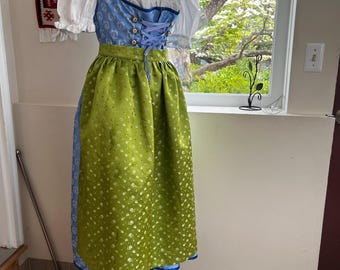 May include: A traditional German dirndl dress displayed on a mannequin. The dress features a white blouse, a blue bodice with a lace-up front, and a green skirt with a floral pattern. The dress is trimmed with blue and white accents.
