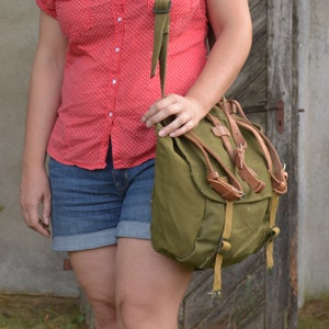 May include: A woman wearing a red and white polka dot shirt and blue denim shorts is carrying a green canvas backpack with brown leather straps.