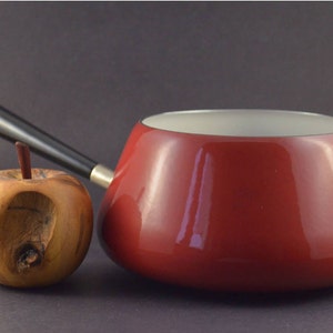 Może przedstawiać: Czerwony emaliowany garnek do fondue z czarnym uchwytem i białym wnętrzem. Garnek stoi na stole obok drewnianego jabłka.