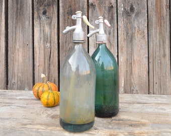 Pode incluir: Dois sifões de soda vintage de vidro com topos e bicos de metal, um transparente e outro verde, sobre uma superfície de madeira com duas pequenas abóboras laranja.