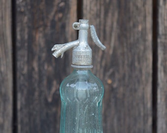 Pode incluir: Uma garrafa de vidro vintage com um sifão de soda de metal. A garrafa é transparente e tem uma superfície texturizada. O sifão é prateado e tem o texto "Putz Kafe" nele.