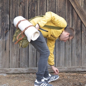 May include: A person wearing a yellow jacket and jeans is carrying a green canvas backpack with a white rolled-up blanket strapped to it.