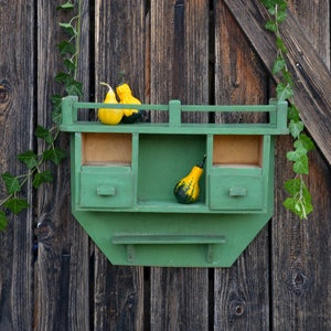 May include: A green wooden shelf with three drawers and a small shelf on the bottom. The shelf is mounted on a wooden wall and has three gourds on the top shelf.