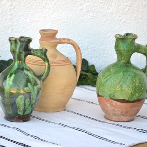 Puede incluir: Tres jarras de cerámica con esmalte verde y marrón. Las jarras tienen un cuerpo redondeado y un asa. Las jarras están sobre un mantel a rayas blancas y negras.