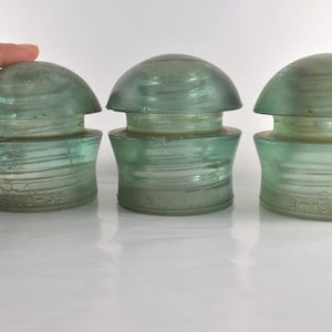 Op de afbeelding: Drie vintage glazen isolatoren in een lichtgroene kleur. De isolatoren zijn koepelvormig met een cilindrische basis. De basis van elke isolator heeft een verhoogde inscriptie met de tekst "5-1/2" en "6 TAS 6-144".