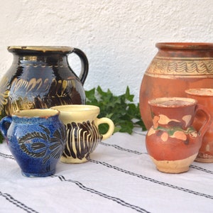 May include: A collection of five hand-painted pottery jugs in various sizes and colors. The jugs are decorated with intricate patterns and designs. The jugs are arranged on a white and black striped tablecloth.