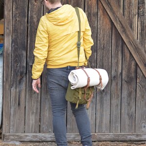 May include: A person wearing a yellow jacket and blue jeans is walking away from a wooden door. They are carrying a green canvas bag with a white blanket inside.