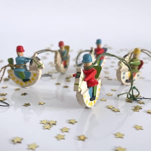 Puede incluir: Adornos navideños de madera con figuras pintadas montadas en caballitos de madera. Las figuras visten trajes rojos, azules y verdes. Los caballitos son blancos con detalles amarillos y verdes. Confeti dorado en forma de estrella.