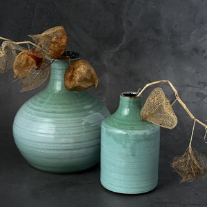 May include: Two green ceramic vases with dried, delicate, brown flowers. The larger vase is round and the smaller vase is cylindrical.