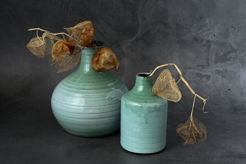 May include: Two green ceramic vases with dried, delicate, brown flowers. The larger vase is round and the smaller vase is cylindrical.