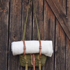May include: A green canvas bag with brown leather straps and buckles hangs on a wooden wall. A white blanket is rolled up and secured inside the bag.
