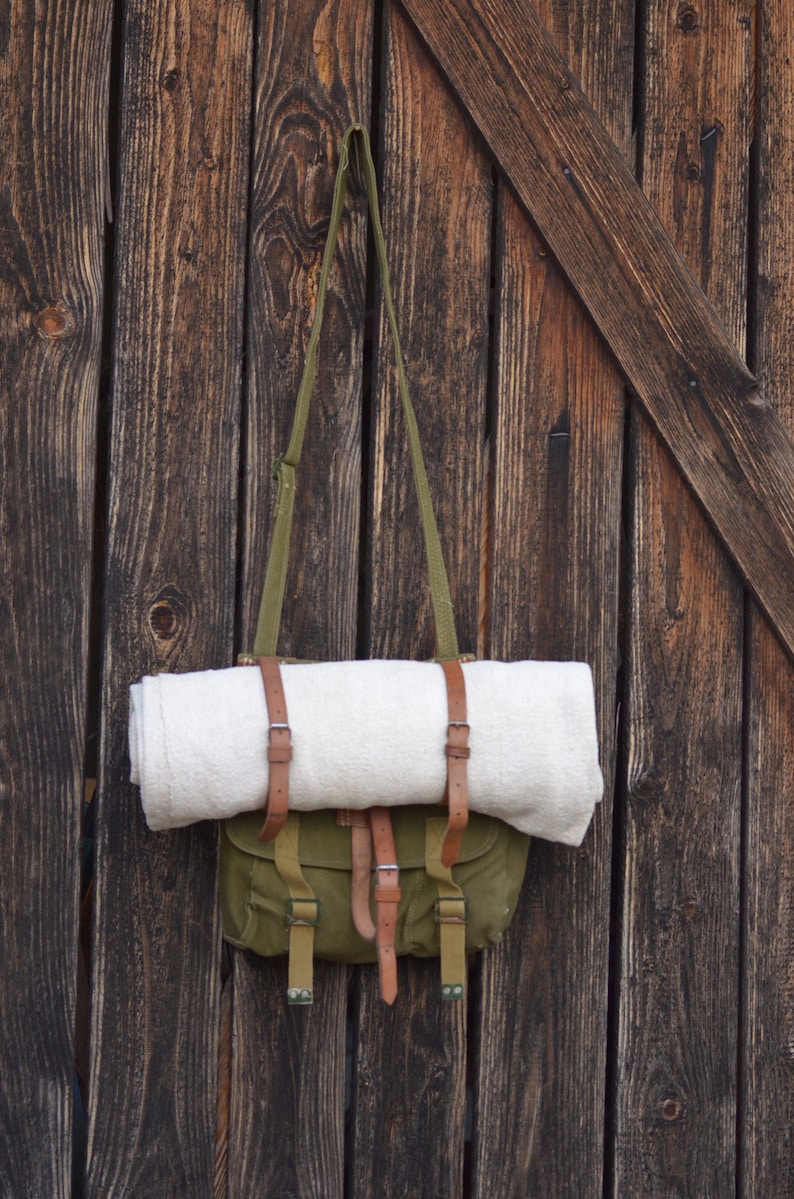 May include: A green canvas bag with brown leather straps and buckles hangs on a wooden wall. A white blanket is rolled up and secured inside the bag.