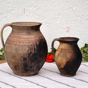 Peut inclure: Deux cruches en terre cuite brunes avec des anses. La plus grande cruche a un motif géométrique rouge et blanc. La plus petite cruche a une seule bande rouge.